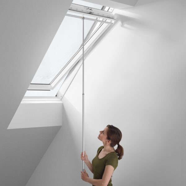 A woman using a silver VELUX telescopic opening rod to open a high-reach center-pivot roof window.