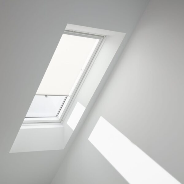 Interior view of a white VELUX roller blind partially extended to filter light in a clean, minimalist white attic room.